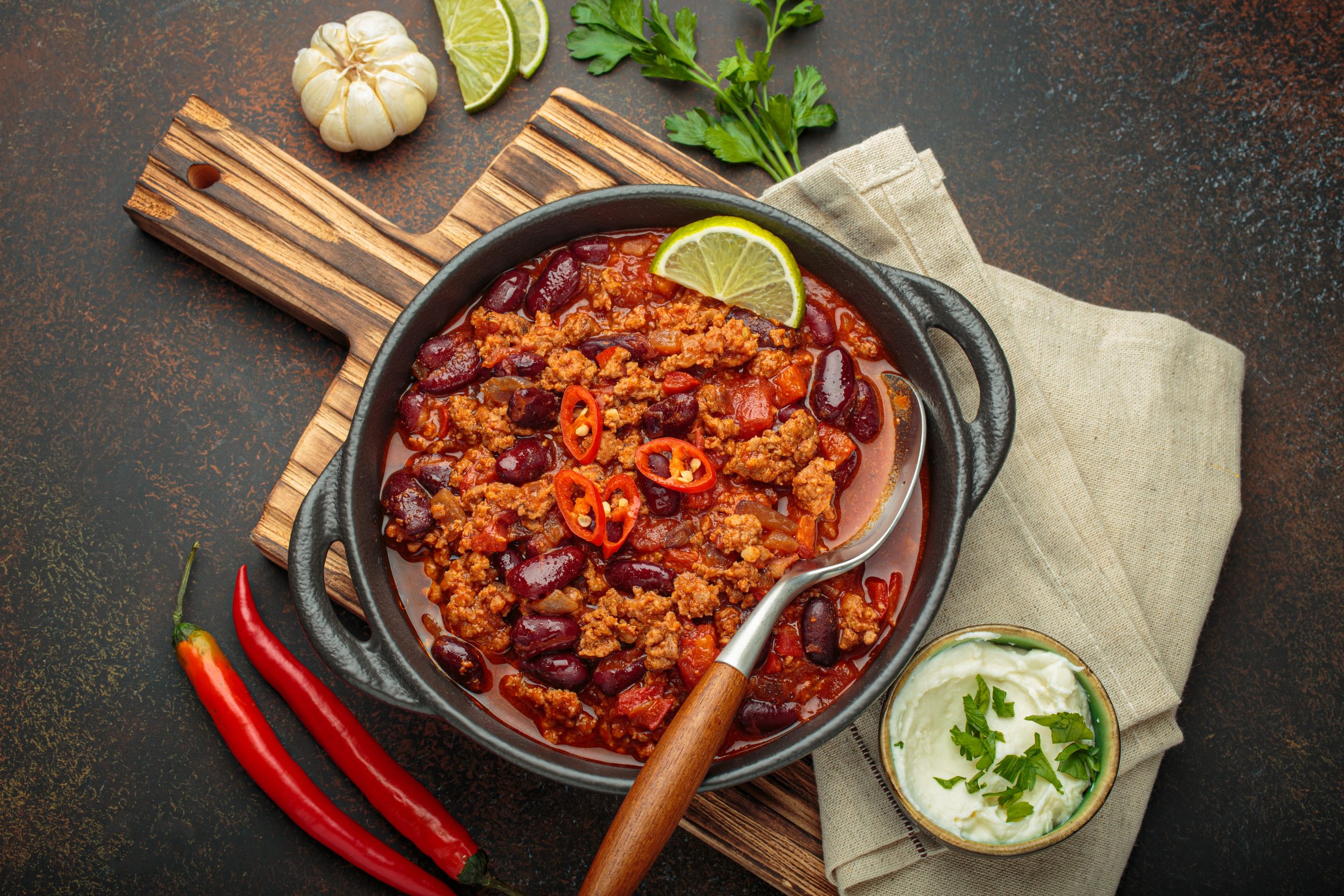 Traditional mexican chili con carne served in a cast iron pot, garnished with lime and red chili peppers, alongside sour cream and fresh ingredients on a rustic wooden board
